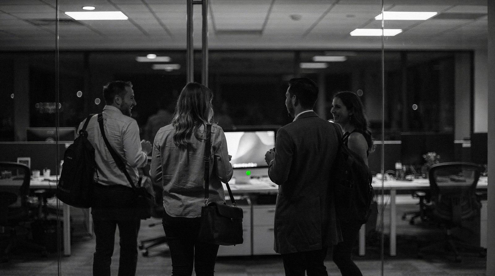 A team leaves the office for the evening, jackets on, laughing; behind them one monitor stays on with a calm green status light