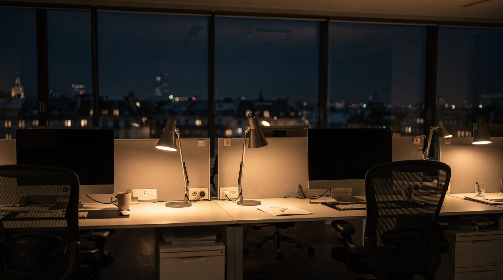 An empty office at night: warm desk lamps on, chairs pushed in, monitors dark, city lights in the windows